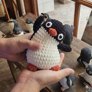 Puede incluir: Una muñeca de pingüino de ganchillo hecha a mano con cuerpo blanco, cabeza y alas negras y pico y pies naranjas. El pingüino está sostenido en las manos de alguien, con otras figuras de pingüinos en el fondo.