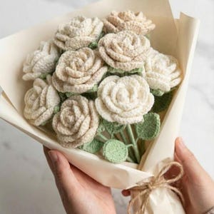 Peut inclure: Un bouquet de roses crochetées crème et beige, avec des feuilles vertes. Les fleurs sont enveloppées dans du papier blanc cassé et attachées avec de la ficelle. Le bouquet fait main est tenu par une personne.