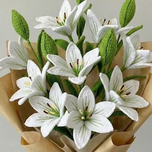 Könnte beinhalten: Ein Strauß handgehäkelter Lilien. Die weißen Blüten haben detaillierte Blütenblätter und braune Staubgefäße, mit grünen Blättern und Knospen. Der Strauß ist in braunes Papier gewickelt und mit Bindfaden gebunden.
