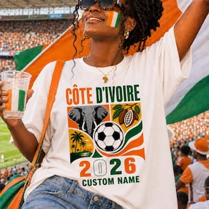May include: A white t-shirt featuring a graphic with the text CÔTE D'IVOIRE and 2026 CUSTOM NAME. The design includes an elephant head, a soccer ball, palm trees, and cocoa pods in orange, green, and black colors. The shirt has a crew neckline.