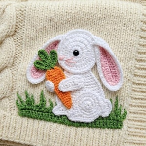May include: A cream-colored knitted square featuring a white crochet bunny holding an orange carrot. The bunny has pink inner ears and black eyes. Green crochet grass is at the bottom. The design is on a textured background.