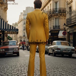 May include: A yellow suit with a tailored jacket and flared pants. The suit is worn by a person standing on a cobblestone street. The image is taken in a European city with vintage cars and buildings.