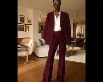 Pode incluir: Um fato de veludo cotelê bordô, composto por um blazer e calças de boca de sino, combinado com uma camisa branca abotoada. O fato apresenta um design clássico de blazer de dois botões. A modelo usa óculos de sol e sapatos claros.