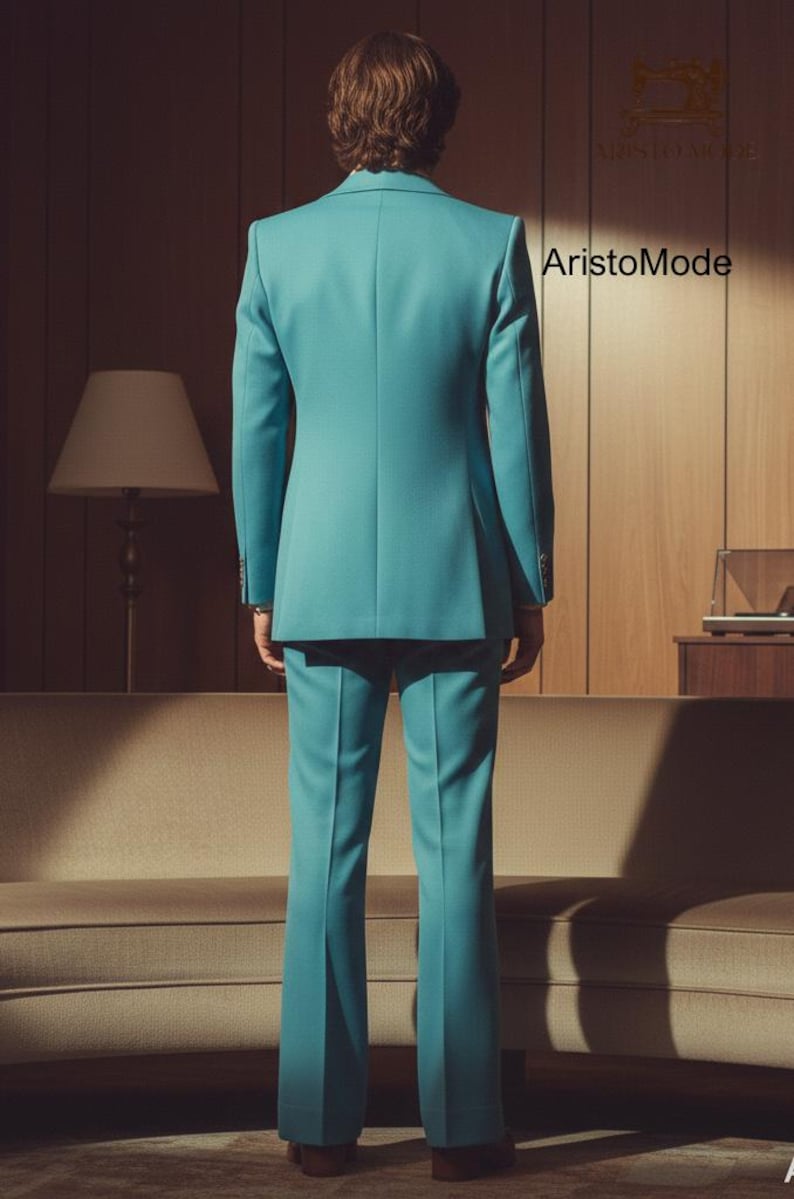 May include: A teal suit, consisting of a blazer and flared trousers, is displayed from the back. The suit is tailored and has a classic design. The brand name "AristoMode" is visible in the background. The setting is a room with a sofa and a lamp.
