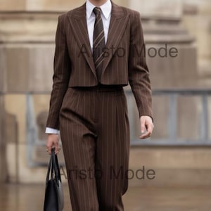 Può includere: Un completo marrone gessato con un blazer corto, pantaloni abbinati e una camicia bianca. Il completo è indossato con una cravatta a righe e occhiali da sole neri. Il marchio "Aristo Mode" è visibile.