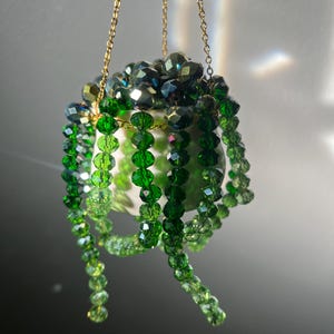 May include: A hanging planter with a white ceramic pot, adorned with strands of faceted green beads. The beads cascade down, creating a decorative effect. The planter is suspended by a delicate gold chain.