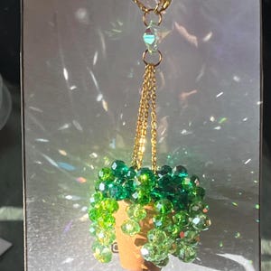 May include: A gold-colored necklace with a pendant shaped like a small potted plant. The plant is made of green beads, and the pot is a light brown color. The necklace has the text "Sweet as Shuga Creations" and "Thank you".