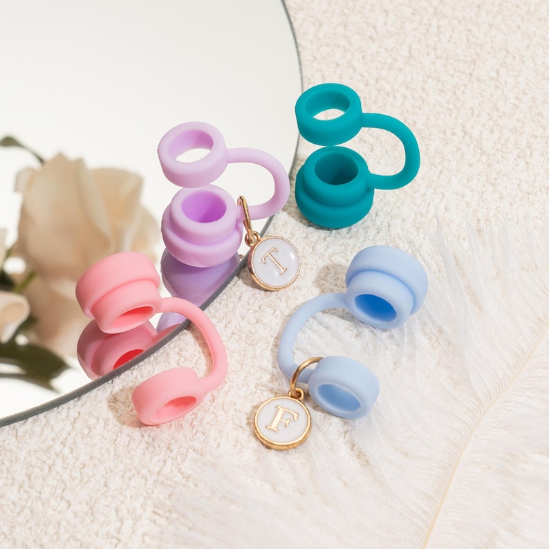 May include: A collection of colorful silicone bottle stoppers in shades of pink, purple, teal, and blue. Each stopper features a loop and a charm with a letter. The stoppers are arranged on a reflective surface.