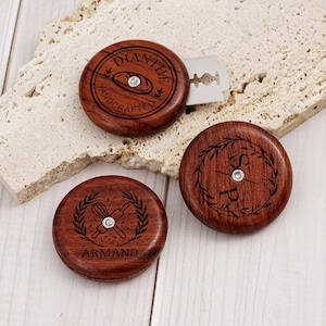 Pode incluir: Três lâminas de pão redondas de madeira com lâminas de metal. Uma apresenta o texto DIANTHE HANDCRAFTED com um ícone de pão, outra as letras SP dentro de uma coroa de louros, e a terceira o nome ARMAND com um ícone de batedor e rolo.