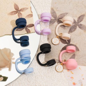 May include: A collection of colorful bottle stoppers in shades of blue, purple, black, and pink. Some stoppers have a small, round charm with a letter. The stoppers are arranged on a reflective surface and a patterned background.