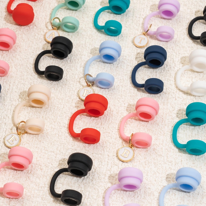 May include: A collection of colorful silicone bottle stoppers in various shades, including red, pink, blue, and black. Each stopper features a curved design and a small, round charm with a letter. The stoppers are arranged on a white, textured surface.