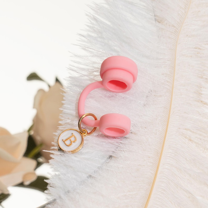 May include: A light pink water bottle cap with a matching tether and a gold-colored charm. The charm has a white background with the letter "B" in gold. The cap is made of a soft, flexible material.