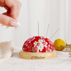 Pode incluir: Uma almofada de alfinetes em forma de cúpula com uma capa de tecido vermelho com um padrão floral branco e rosa. A almofada assenta numa base redonda de madeira clara gravada com o nome Adeline. Várias agulhas de costura prateadas estão inseridas.