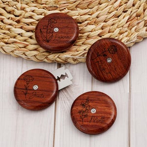 May include: Four circular wooden seam rippers with dark brown wood grain, each featuring a unique engraved floral design and a name. One seam ripper is partially open, revealing a sharp metal blade. The names Laura, Rachel, Jenta, and Miette are engraved.