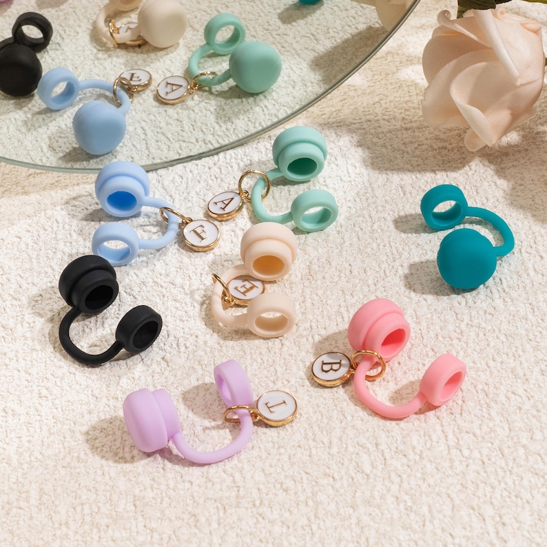 May include: A collection of colorful earbud accessories in various shades, including black, blue, pink, and green. Each accessory features a round, bulbous end and a loop, some with gold-toned initial charms. The items are arranged on a textured, off-white surface.