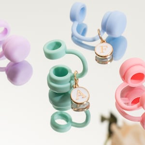 May include: Close-up of several colorful water bottle accessories. The accessories are in shades of pink, green, blue, and purple. Each has a circular top and a curved spout. Gold-colored charms with white enamel and letters hang from some of the accessories.