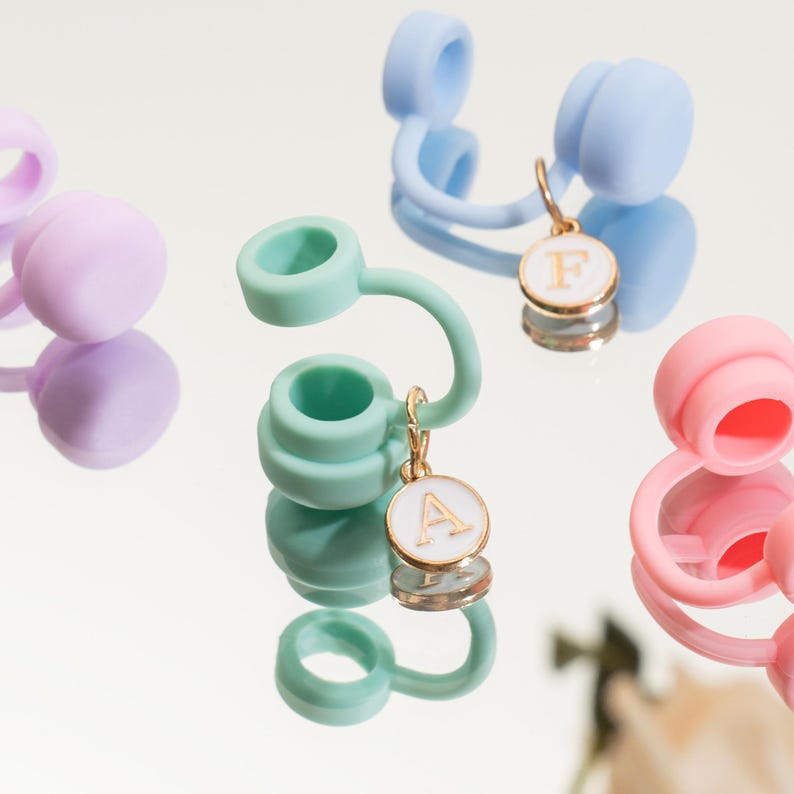 May include: Close-up of several colorful water bottle accessories. The accessories are in shades of pink, green, blue, and purple. Each has a circular top and a curved spout. Gold-colored charms with white enamel and letters hang from some of the accessories.