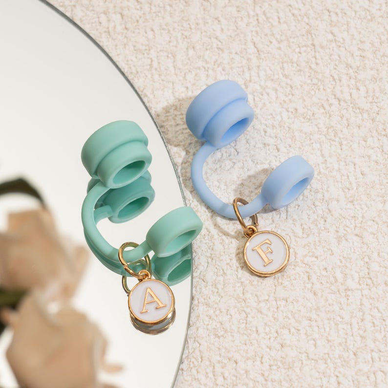 May include: Two silicone phone charms, one mint green and one light blue, each with a gold-colored ring and a white circular charm. The white charms have the letters "A" and "F" in gold. The charms are displayed on a reflective surface.