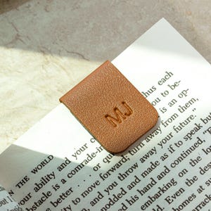 Può includere: Un segnalibro in pelle marrone con le iniziali "MJ" in rilievo è posto sull'angolo di un libro. Il segnalibro è rettangolare con angoli arrotondati. Le pagine del libro sono visibili, mostrando testo stampato.