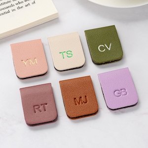 Può includere: Sei segnalibri magnetici in pelle in vari colori: rosa, crema, verde oliva, malva, marrone e lavanda. Ogni segnalibro presenta iniziali in oro o verde, tra cui "YM", "TS", "CV", "RT", "MJ" e "GB".