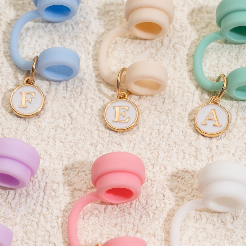 May include: A collection of colorful bottle stoppers with gold-colored initial charms. The stoppers are in various pastel colors, including blue, pink, and green. Each stopper has a circular charm with a letter in the center, such as "F", "E", and "A".