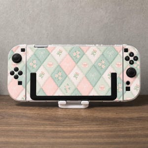 Könnte beinhalten: Eine tragbare Spielkonsole mit einer Schutzfolie in einem pastellfarbenen Rautenmuster in Hellrosa und Mintgrün. Das Design zeigt kleine Illustrationen von weißen Schleifen, blühenden Zweigen und Teetassen auf der gesamten Oberfläche.