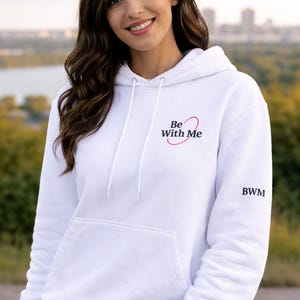 Puede incluir: Sudadera con capucha blanca con bolsillo delantero y cord&oacute;n. El texto "Be With Me" est&aacute; impreso en el pecho con un dise&ntilde;o de coraz&oacute;n rosa. Las letras "BWM" est&aacute;n en la manga.