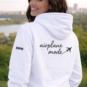 Puede incluir: Sudadera con capucha blanca con el texto "airplane mode" y un peque&ntilde;o gr&aacute;fico de avi&oacute;n. La sudadera tiene las letras "BWM" en la manga izquierda. La persona de la foto est&aacute; sonriendo.