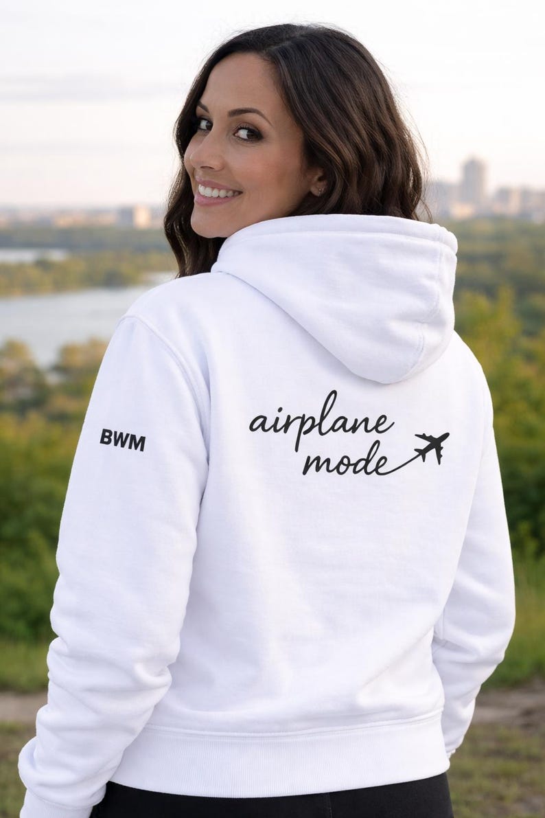 Puede incluir: Sudadera con capucha blanca con el texto "airplane mode" y un peque&ntilde;o gr&aacute;fico de avi&oacute;n. La sudadera tiene las letras "BWM" en la manga izquierda. La persona de la foto est&aacute; sonriendo.
