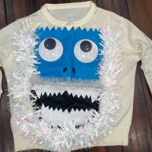 May include: Cream-colored cardigan sweater with a blue monster face design. The monster face has large white eyes, a black mouth, and is surrounded by white tinsel. A fun and festive holiday sweater.