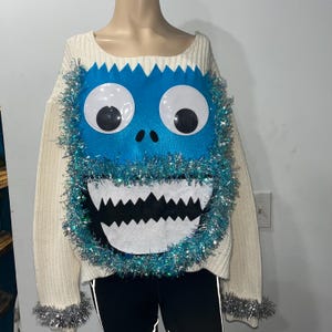 May include: Off-white knit sweater with a large blue monster face design. The monster face features large white eyes with black pupils, a jagged mouth, and a blue felt face. Silver tinsel trims the face and cuffs.
