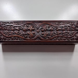 Puede incluir: Una caja rectangular de madera de color marrón oscuro con intrincadas tallas florales simétricas en la superficie superior. La tapa muestra un patrón de relieve detallado con motivos de enredaderas y elementos circulares centrales.