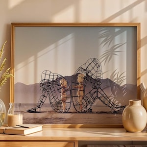 May include: A framed art print featuring a wire sculpture of two figures in a seated pose. The artwork is displayed on a wooden cabinet with decorative vases, a glass vase with dried plants, and books. The color palette is neutral with warm tones.