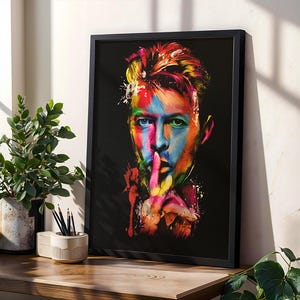 Może przedstawiać: Oprawiony wydruk artystyczny przedstawiający kolorowy portret osoby z palcem na ustach. Grafika wykorzystuje żywą paletę niebieskiego, żółtego, różowego i czerwonego na czarnym tle. Rama jest czarna.