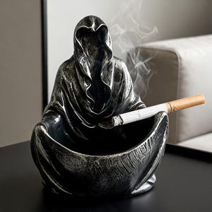 Op de afbeelding: Een donkergrijze asbak in de vorm van een figuur met capuchon, met een brandende sigaret. De asbak heeft een getextureerd oppervlak en geeft rook af. Het ontwerp is gedetailleerd en de algehele esthetiek is gotisch.