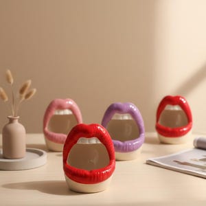 Puede incluir: Cuatro jarrones de cerámica con forma de labios en rojo, rosa y morado. Los jarrones tienen bocas abiertas y dientes blancos. Un pequeño jarrón con flores secas está a la izquierda. Una revista está a la derecha.