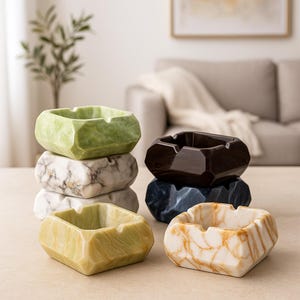 May include: A collection of decorative ashtrays in various colors and patterns. The ashtrays are square-shaped with a faceted design, and are stacked in groups. Colors include green, marble, brown, blue, and cream with gold veining. The ashtrays are made of ceramic.