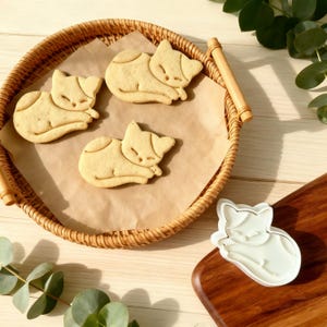 May include: Three cat-shaped biscuits sit on parchment paper in a wicker basket. The biscuits are light brown with darker markings. A white cat-shaped biscuit cutter is on a wooden board.