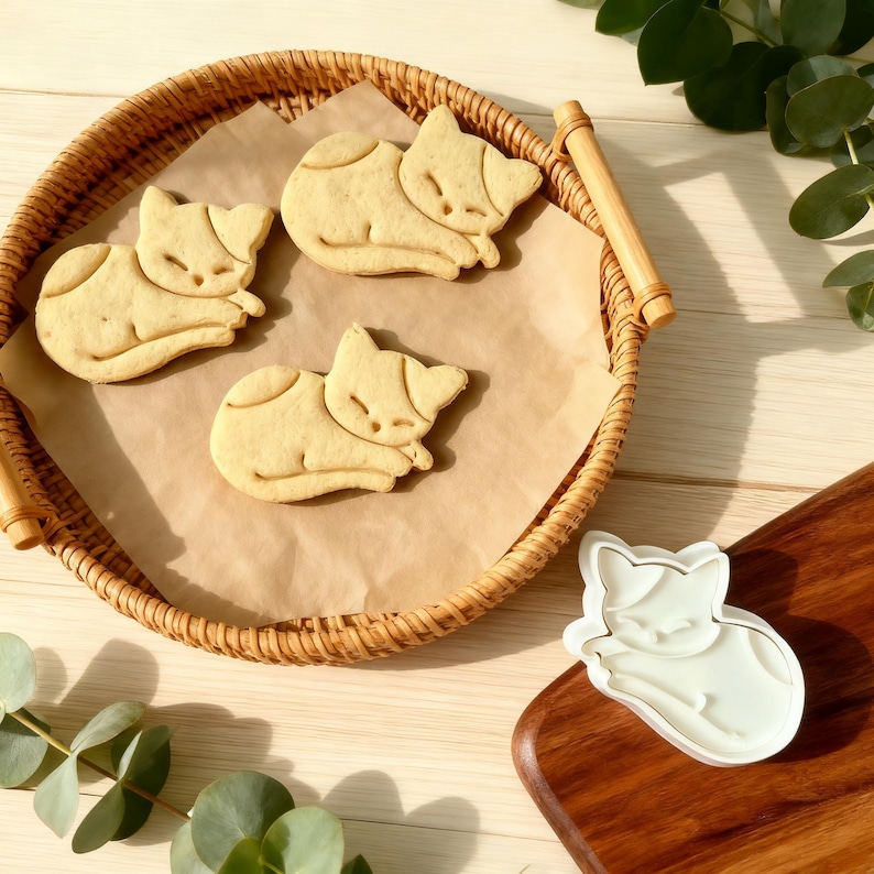 May include: Three cat-shaped biscuits sit on parchment paper in a wicker basket. The biscuits are light brown with darker markings. A white cat-shaped biscuit cutter is on a wooden board.