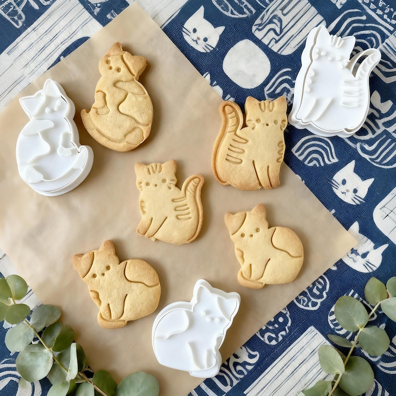 May include: A selection of cat-shaped biscuits and white plastic cookie cutters are arranged on a brown surface. The biscuits are light brown with etched details. The background is a blue and white patterned fabric with some green leaves.