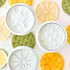 Puede incluir: Vista cenital de cortadores de galletas blancos con diseños de limón y varias galletas con forma de limón. Las galletas son amarillas, verdes y blancas, con texturas detalladas. Los cortadores tienen diferentes patrones, incluyendo rodajas de limón y limones enteros.