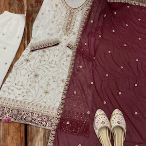 May include: An ivory-colored embroidered salwar kameez set with matching pants and a maroon dupatta. The set features gold and maroon embellishments, with a pair of embroidered slip-on shoes. The outfit is displayed on a wooden background.