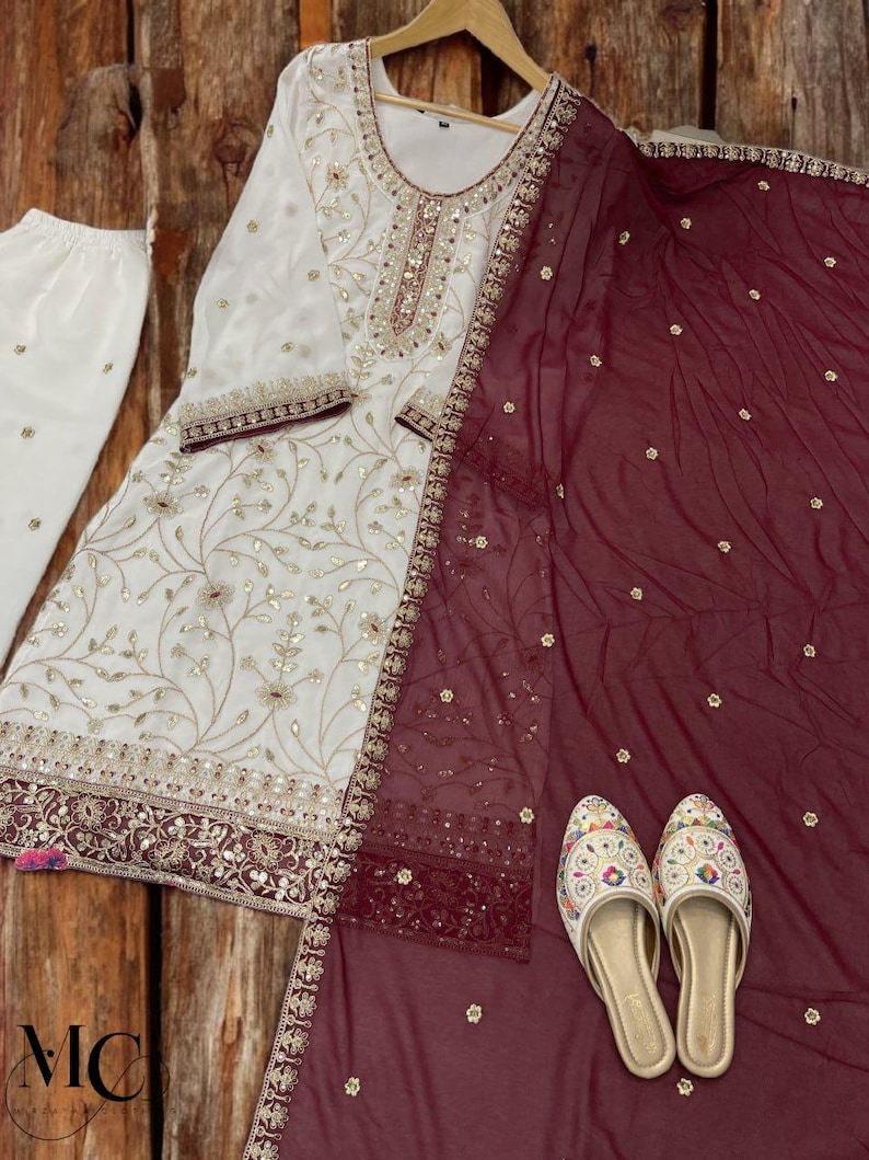 May include: An ivory-colored embroidered salwar kameez set with matching pants and a maroon dupatta. The set features gold and maroon embellishments, with a pair of embroidered slip-on shoes. The outfit is displayed on a wooden background.