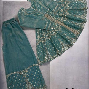 Pu&ograve; includere: Completo formale color teal con top a maniche lunghe e pantaloni abbinati. L'ensemble &egrave; impreziosito da dettagli intricati di paillettes e perline argentate. L'outfit &egrave; esposto su una superficie grigio chiaro.
