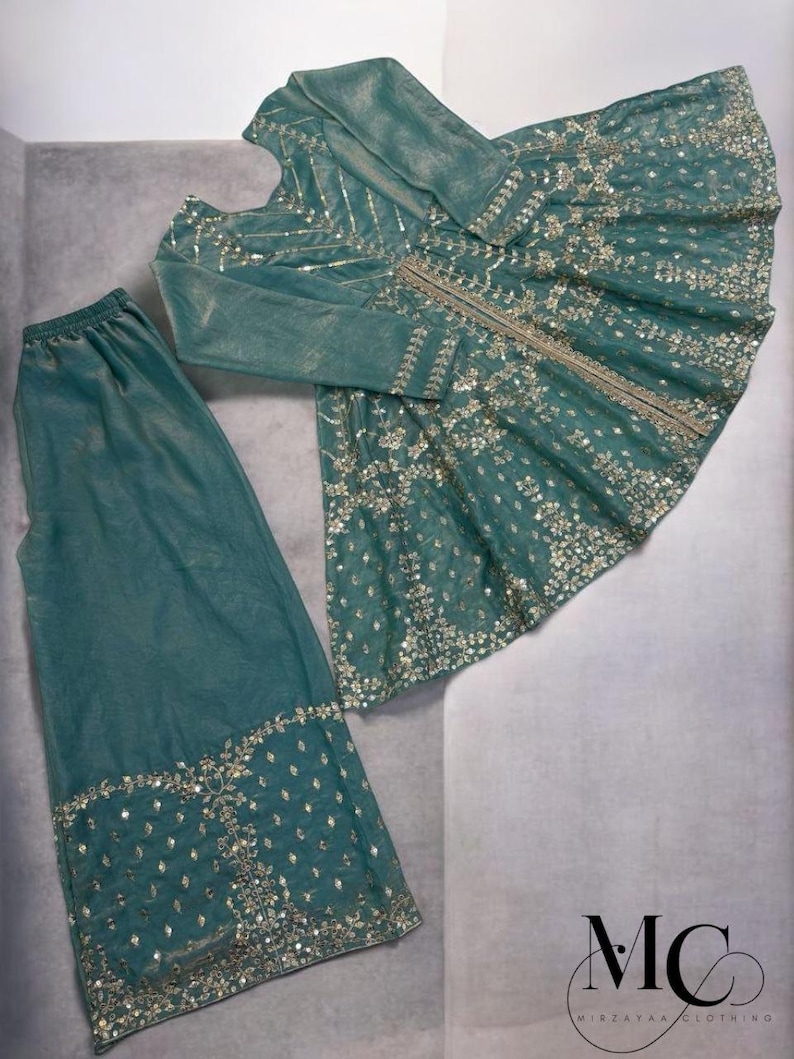 Pu&ograve; includere: Completo formale color teal con top a maniche lunghe e pantaloni abbinati. L'ensemble &egrave; impreziosito da dettagli intricati di paillettes e perline argentate. L'outfit &egrave; esposto su una superficie grigio chiaro.
