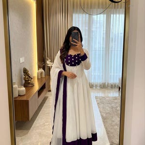 May include: A white dress with a purple bodice and trim, paired with a matching scarf. The dress features long sleeves and a flowing skirt. The outfit is captured in a mirror, reflecting the interior of a well-lit room.