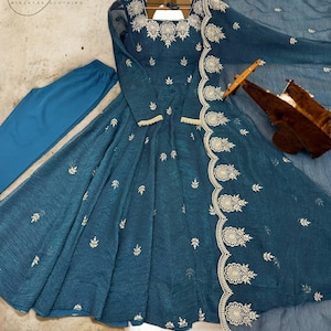 Puede incluir: Un vestido de color turquesa con mangas largas y bordados dorados intrincados en el escote, las mangas y el dobladillo. El vestido tiene una falda fluida y se combina con pantalones turquesa a juego y una bufanda transparente. El vestido está colgado en una percha de madera.