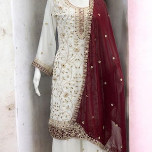 May include: White embroidered salwar kameez with palazzo pants and a maroon dupatta. The ensemble features intricate gold embroidery on the neckline, sleeves, and hem. The mannequin has a gold-colored head.