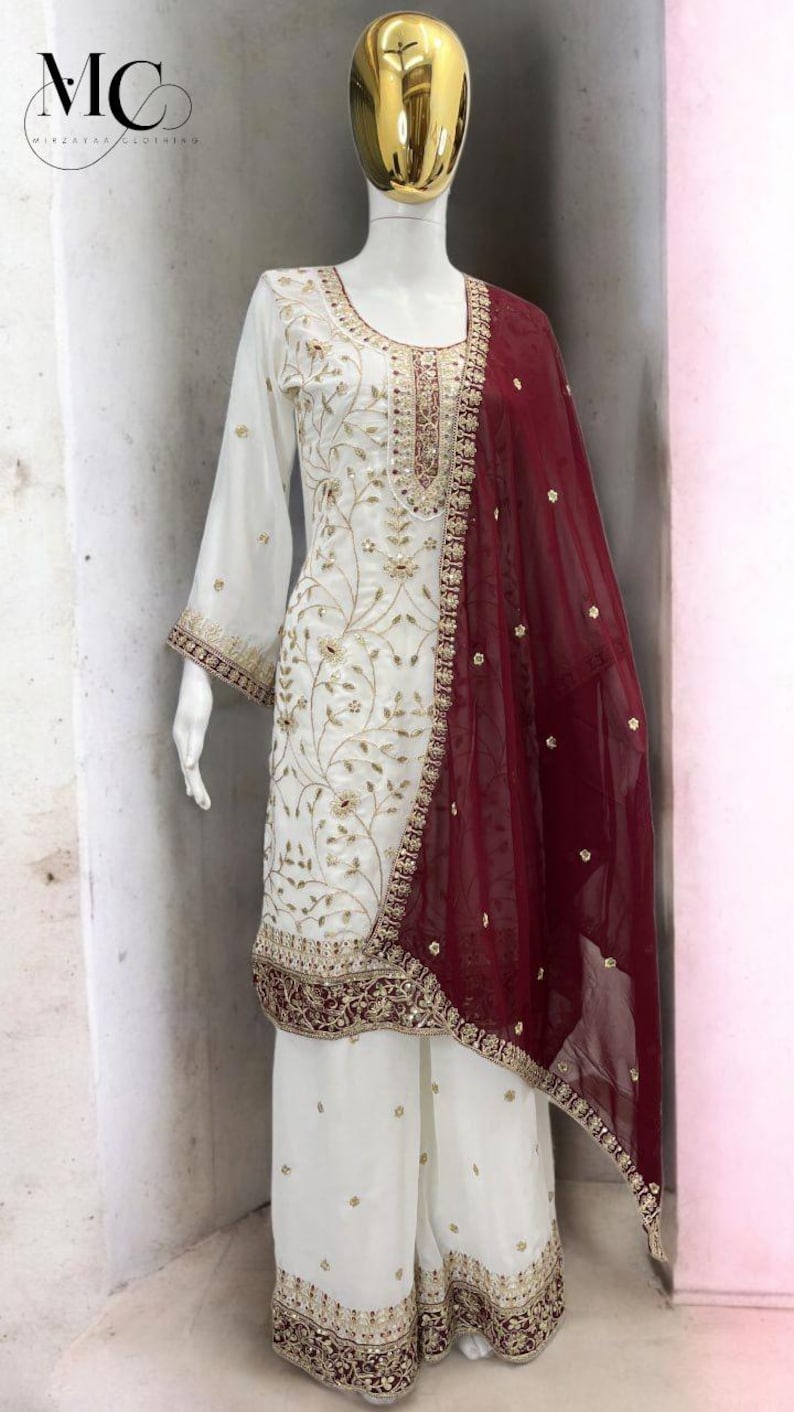 May include: White embroidered salwar kameez with palazzo pants and a maroon dupatta. The ensemble features intricate gold embroidery on the neckline, sleeves, and hem. The mannequin has a gold-colored head.