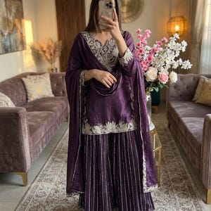 Może przedstawiać: Głęboki fioletowy zestaw salwar kameez z misternym srebrnym haftem na dekolcie, rękawach i obszyciu. Zestaw zawiera pasującą dupattę i spodnie z szerokimi nogawkami. Str&oacute;j wykonany jest z błyszczącej tkaniny.